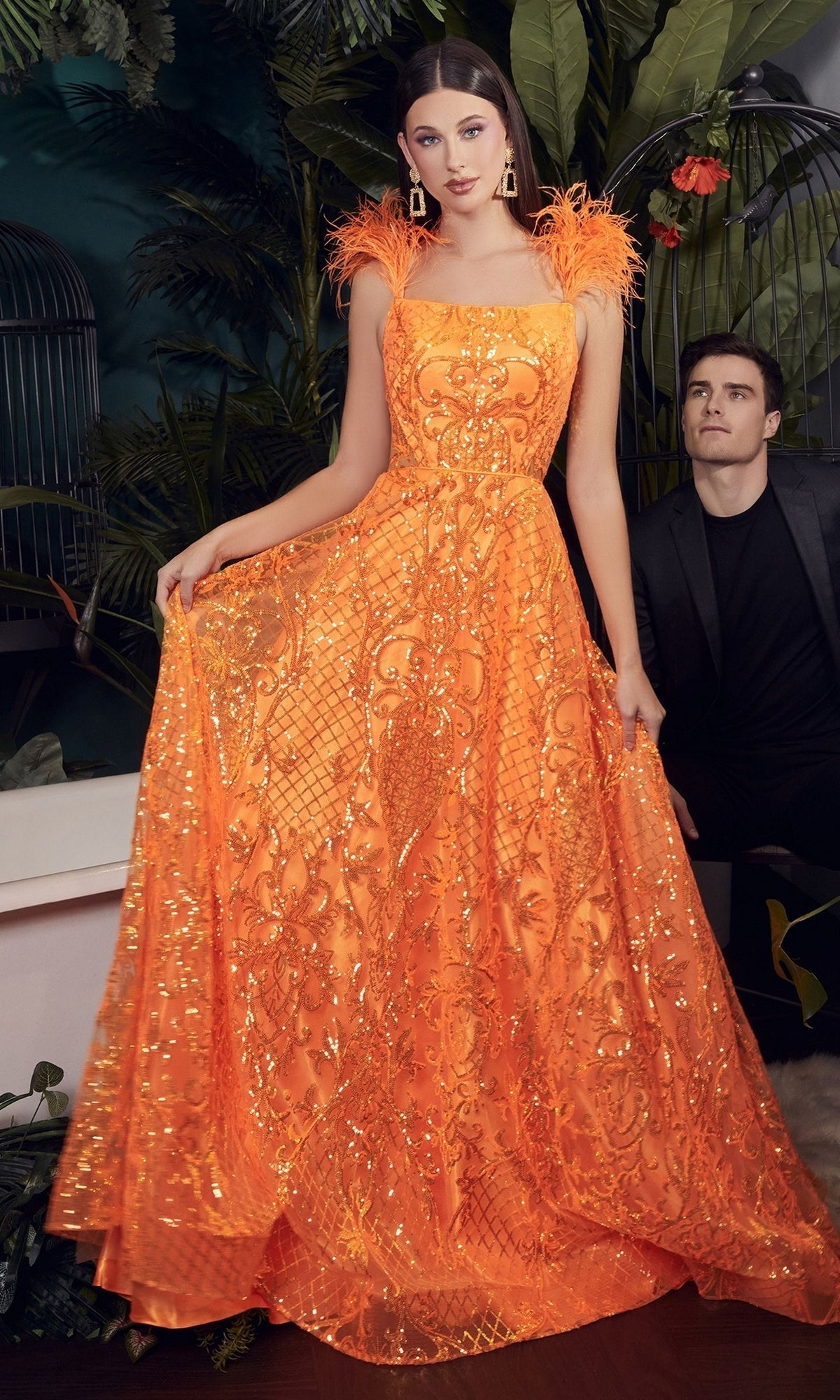 A woman wears the Feathered Neon Orange Long Sequin Prom Dress KV1076 with feathered shoulder straps among plants, while a man in a black suit sits nearby in the background.